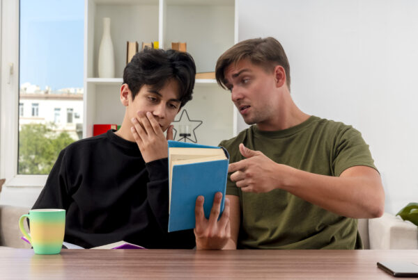 confident young blonde handsome man holding and pointing at book sitting at table and looking at young brunette handsome guy putting hand on mouth looking at book inside design living room